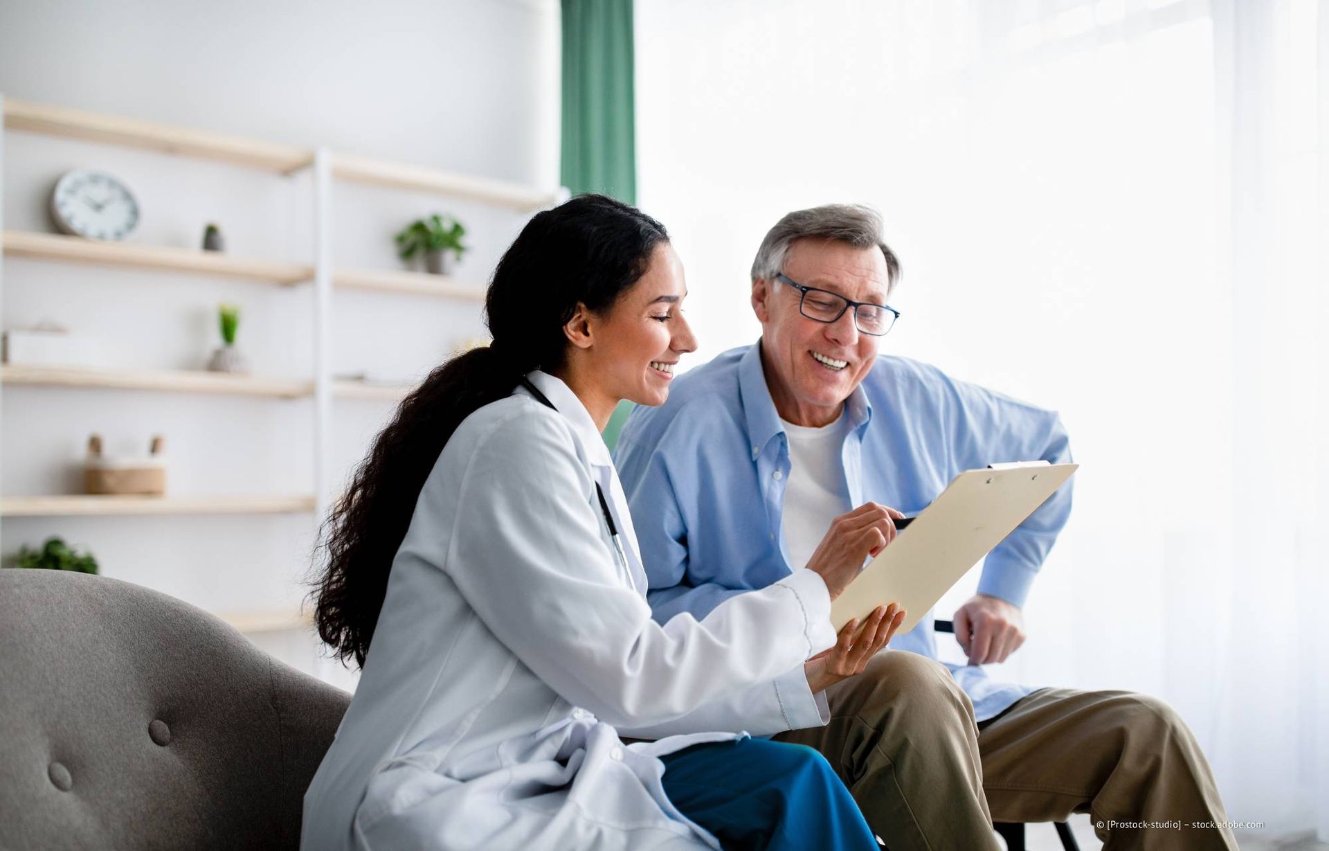 Ein Arzt zeigt einem Patienten eine Patientenakte; beide lächeln in einem hell erleuchteten Raum.