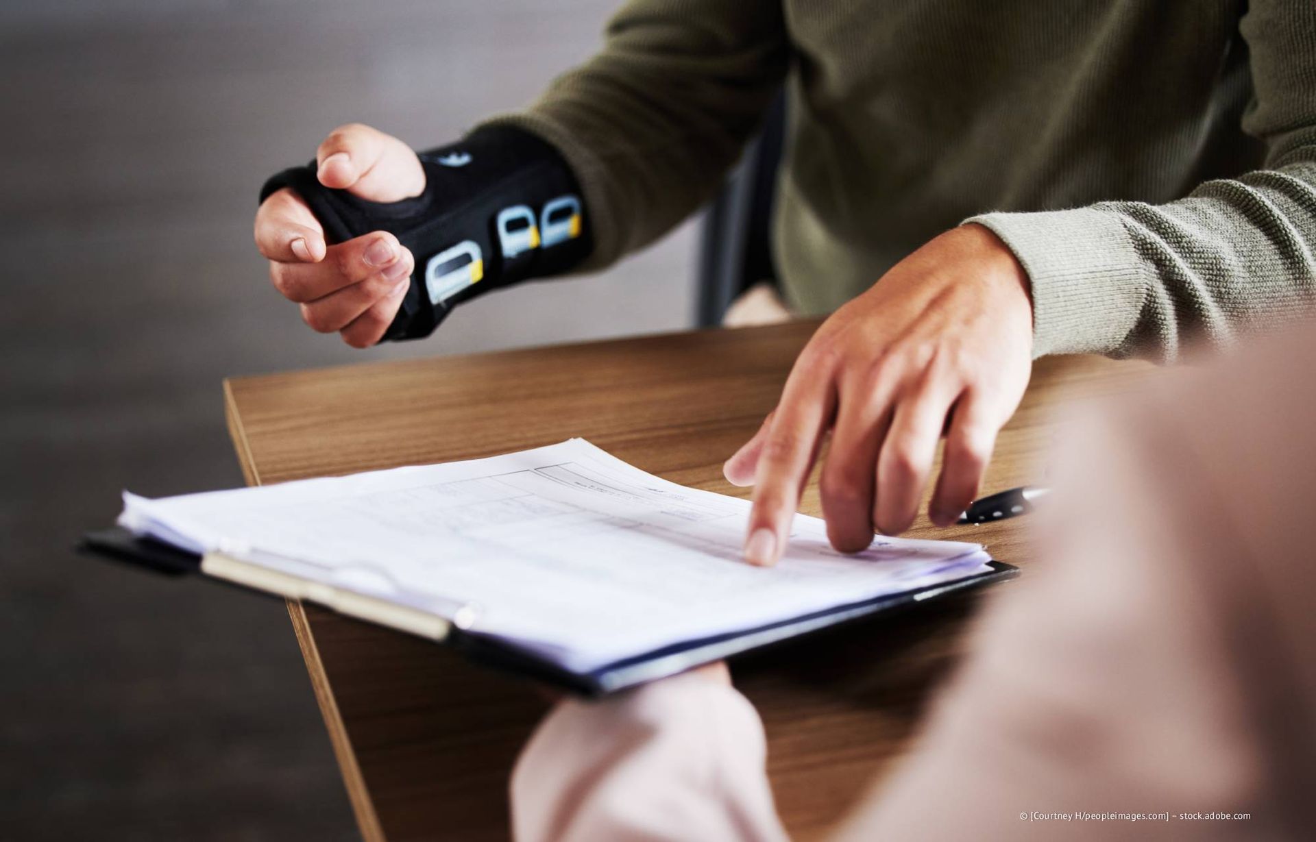 Eine Person, die ein Gerät am Handgelenk trägt, zeigt während einer Besprechung auf ein Dokument auf einem Klemmbrett.