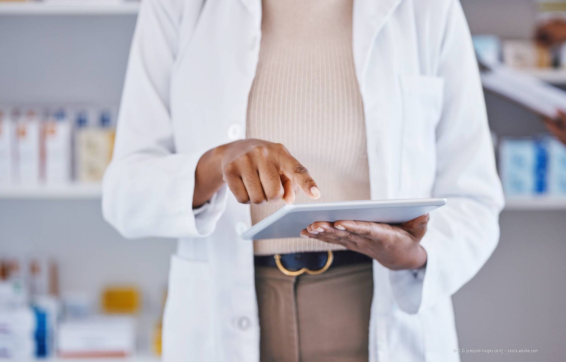 Ein Apotheker benutzt in einer Apotheke eine Tablette; er trägt einen Laborkittel, braune Hosen und ein neutrales Oberteil. Im Hintergrund sind Regale mit Medikamenten zu sehen.