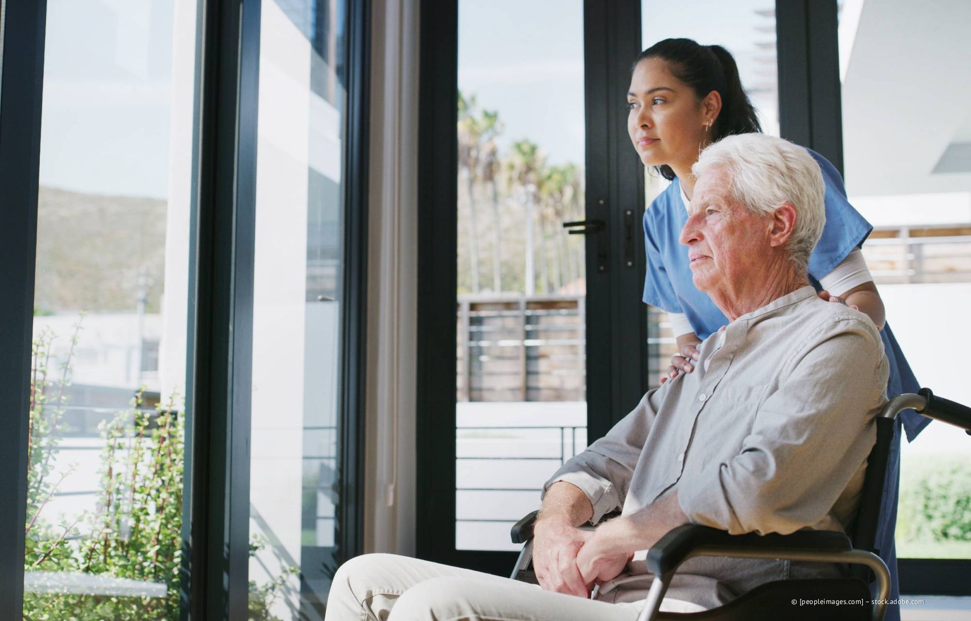 Eine Frau in OP-Kleidung hilft einem Mann im Rollstuhl an einem großen Fenster. Beide schauen hinaus.