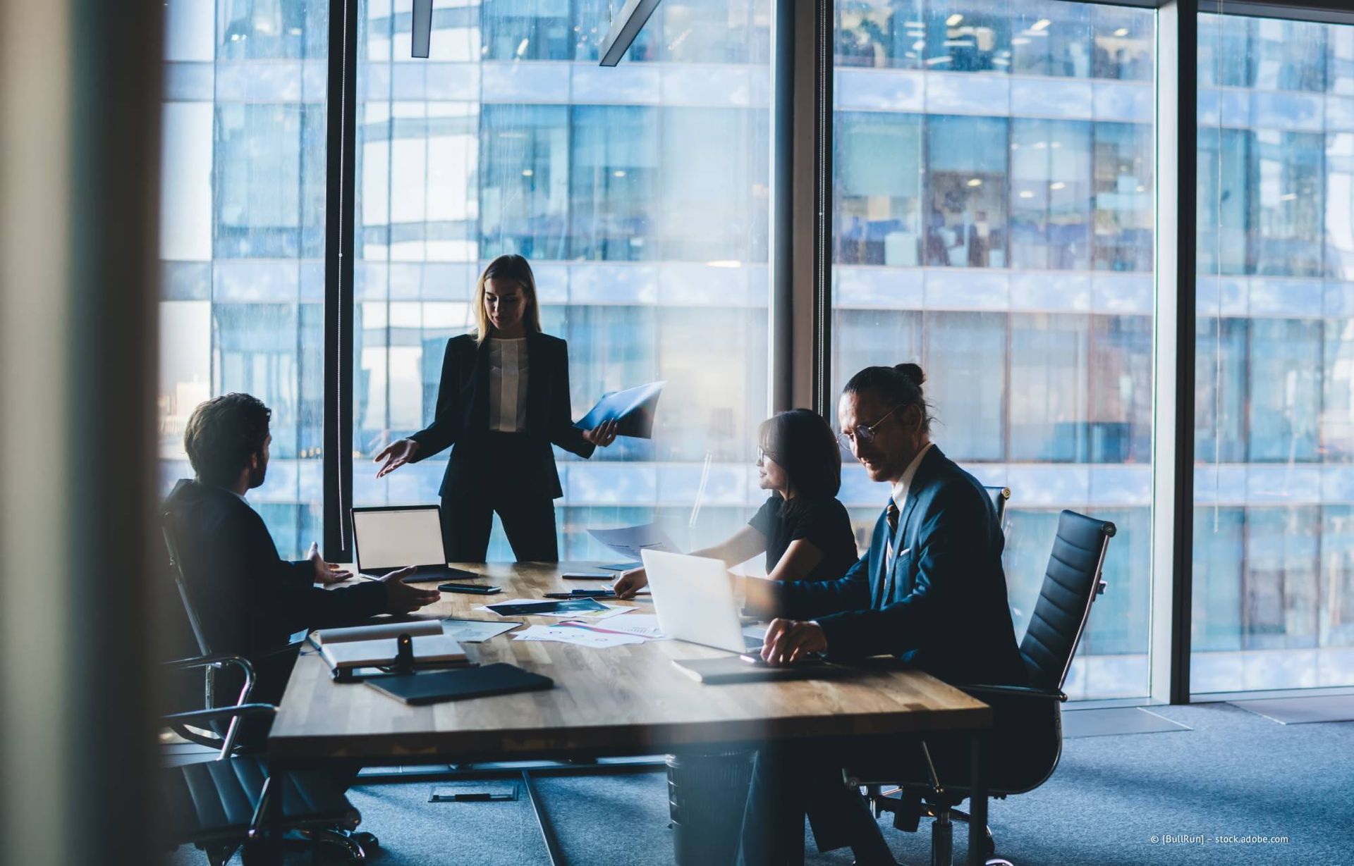 Geschäftstreffen in einem modernen Büro; eine Frau steht und präsentiert ihren am Tisch sitzenden Kollegen.