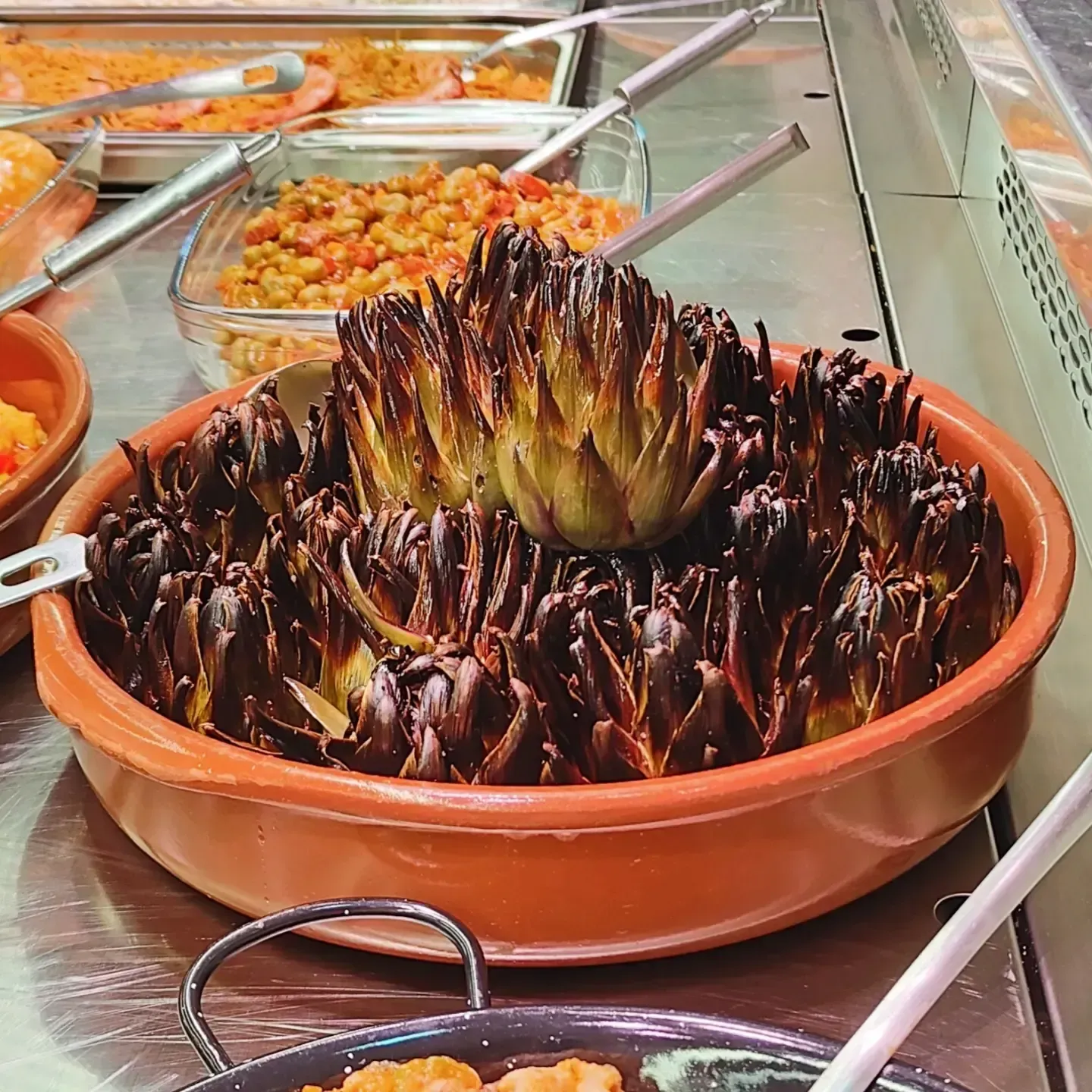 Bandeja de alcachofas a la parrilla en un bufé, con cucharas de metal para servir y otros platos detrás.