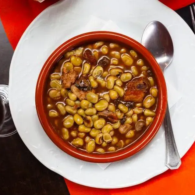 Un tazón de sopa de frijoles con carne en un plato blanco, con una cuchara sobre un mantel rojo.