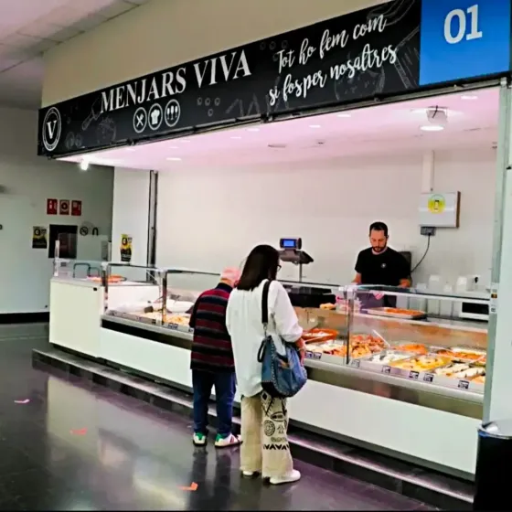 Mostrador de panadería con pasteles, dos clientes y un empleado detrás del mostrador de cristal.