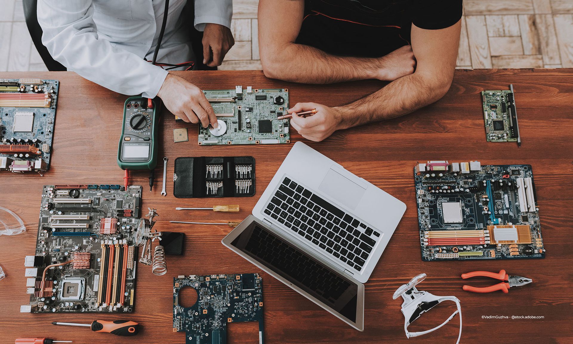 Zwei Personen arbeiten an einem Holztisch mit einem Laptop, Werkzeugen und Komponenten an Computerplatinen.