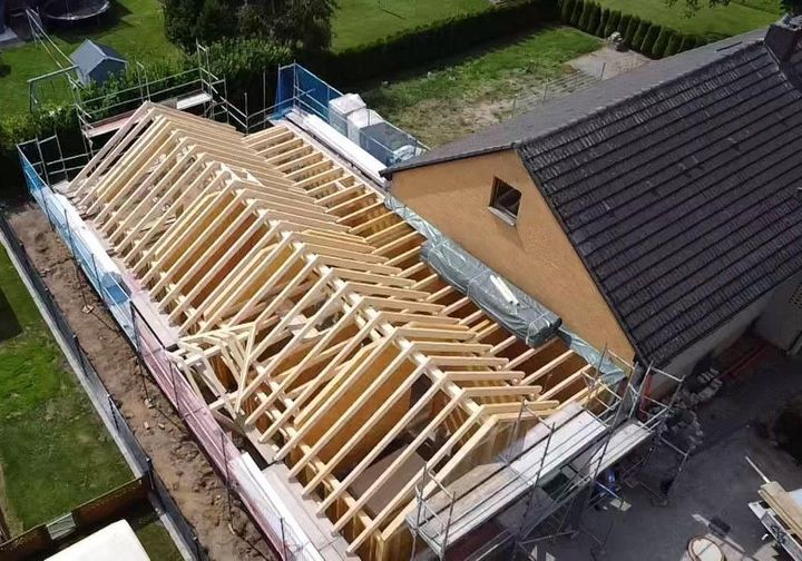 Luftaufnahme einer Baustelle: Auf einem Haus wird ein hölzerner Dachstuhl neben einem bestehenden Dach errichtet.