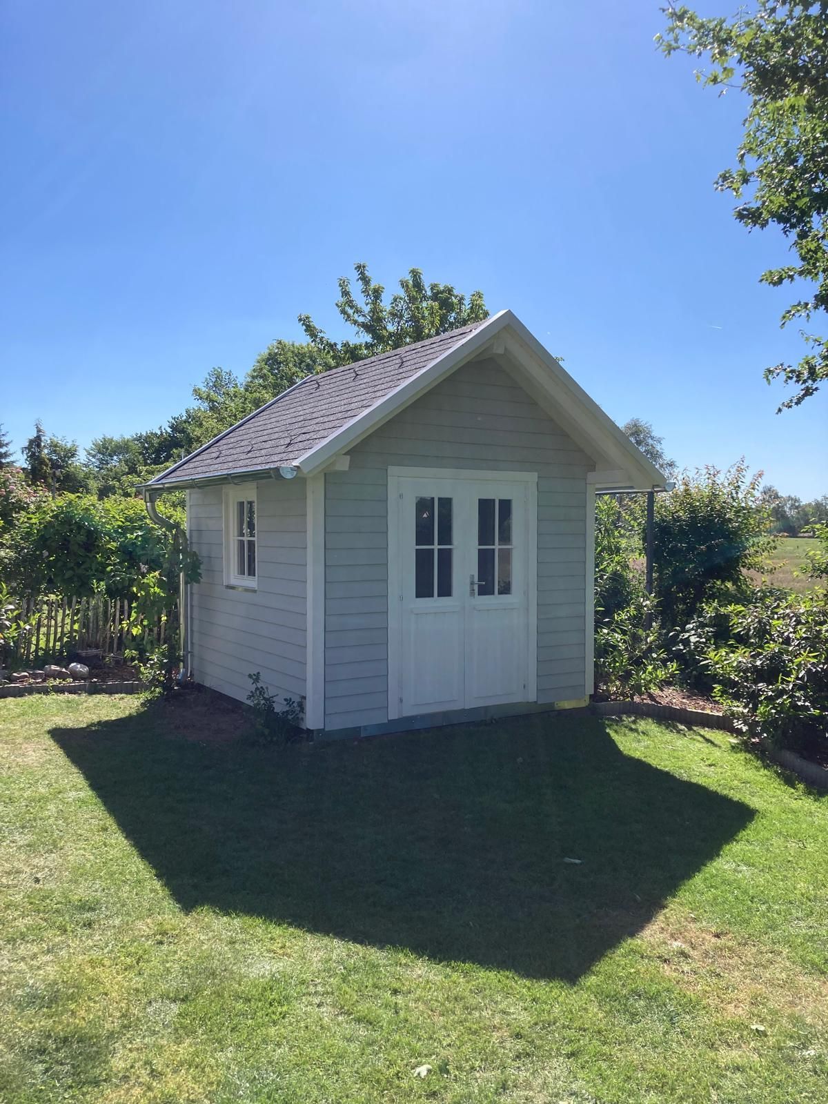 Hellblaues Gartenhaus mit weißen Türen und Fenstern, umgeben von grünem Gras und Laub an einem sonnigen Tag.