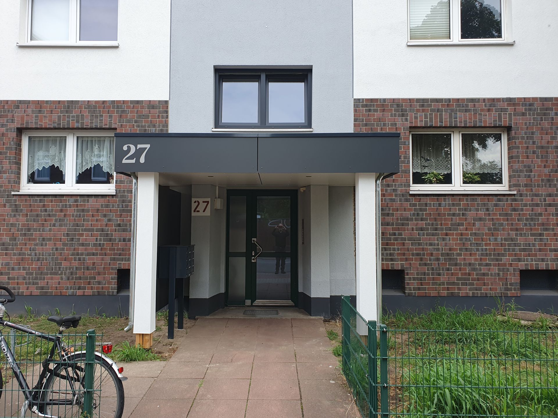 Eingang eines Mehrfamilienhauses, Fassade aus Backstein und Grau, Nummer 27 über der Tür, davor geparktes Fahrrad, Grünanlage.