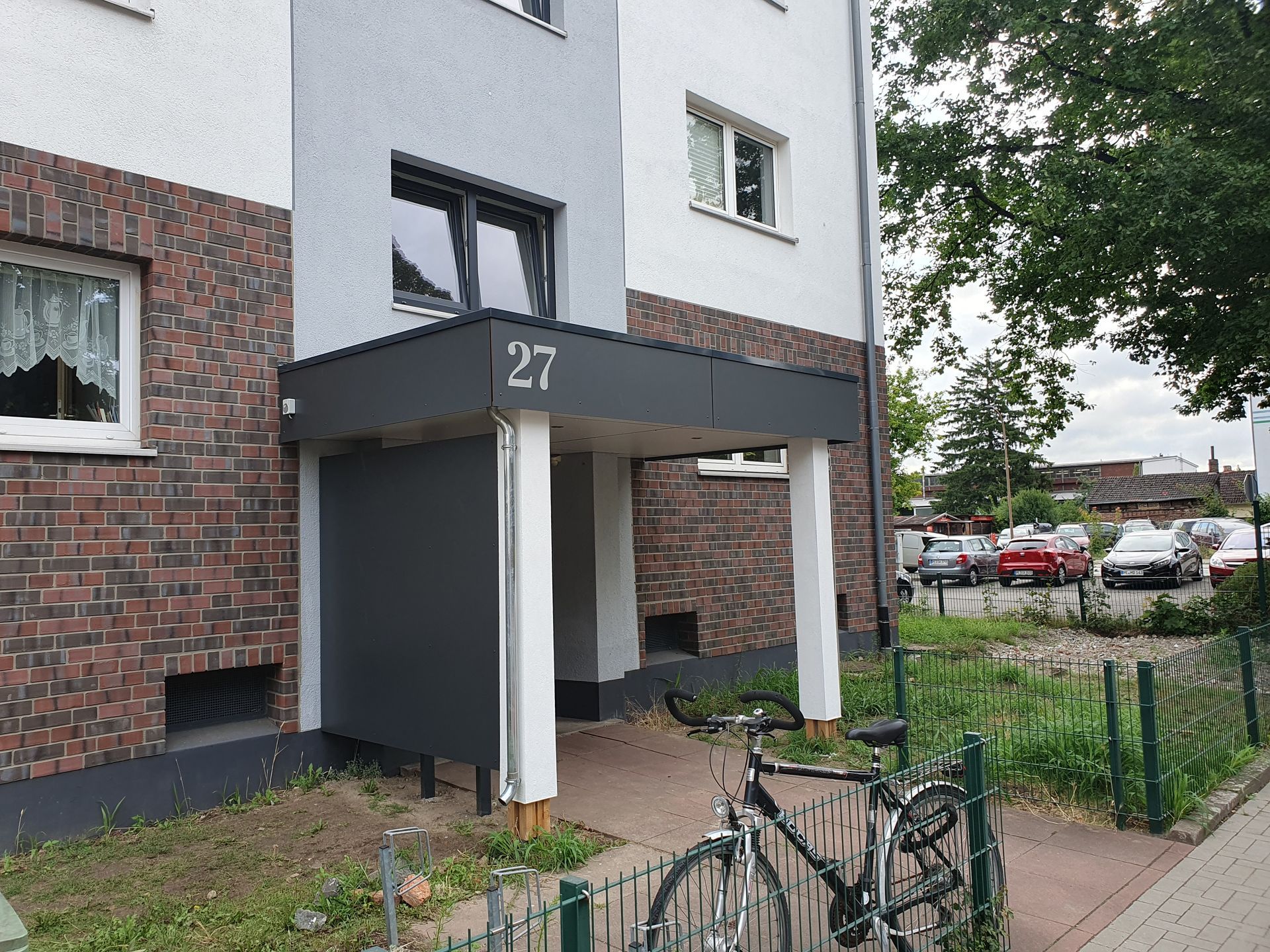 Gebäudeeingang mit der Nummer 27, graue Markise, Ziegel- und weiße Wände, mit sichtbaren geparkten Fahrrädern und Autos.