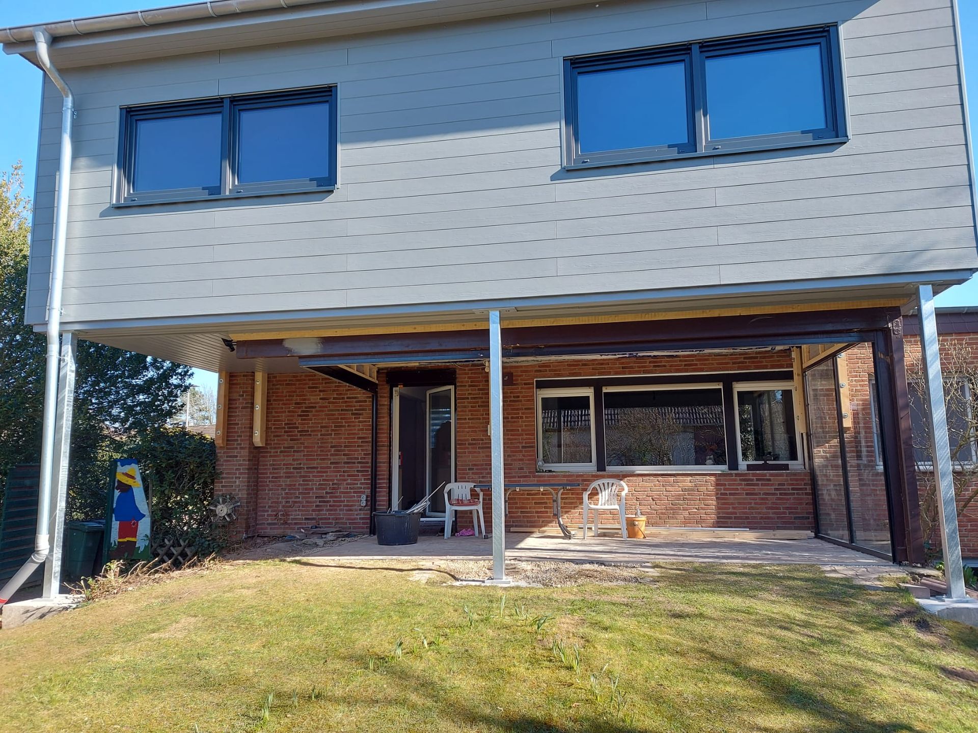 Zweistöckiges Haus mit überdachter Terrasse. Graue Außenverkleidung, Ziegelwände und Rasen.
