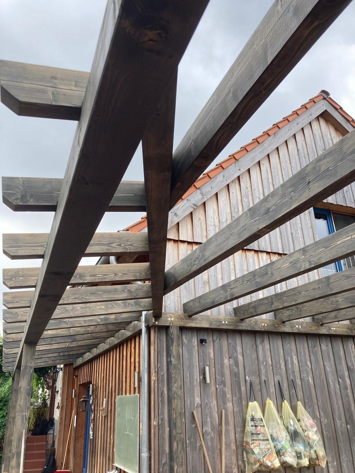 An einem Holzgebäude befestigte Holzpergola unter bewölktem Himmel.