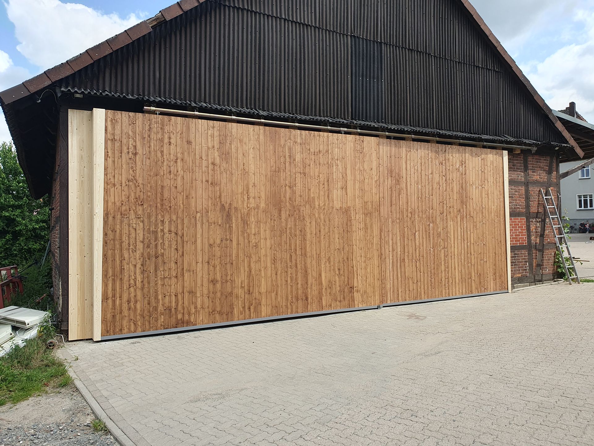 Großes, geschlossenes Scheunentor aus Holz an einem Gebäude mit dunklem Dach und Ziegelfassade. Betonauffahrt.