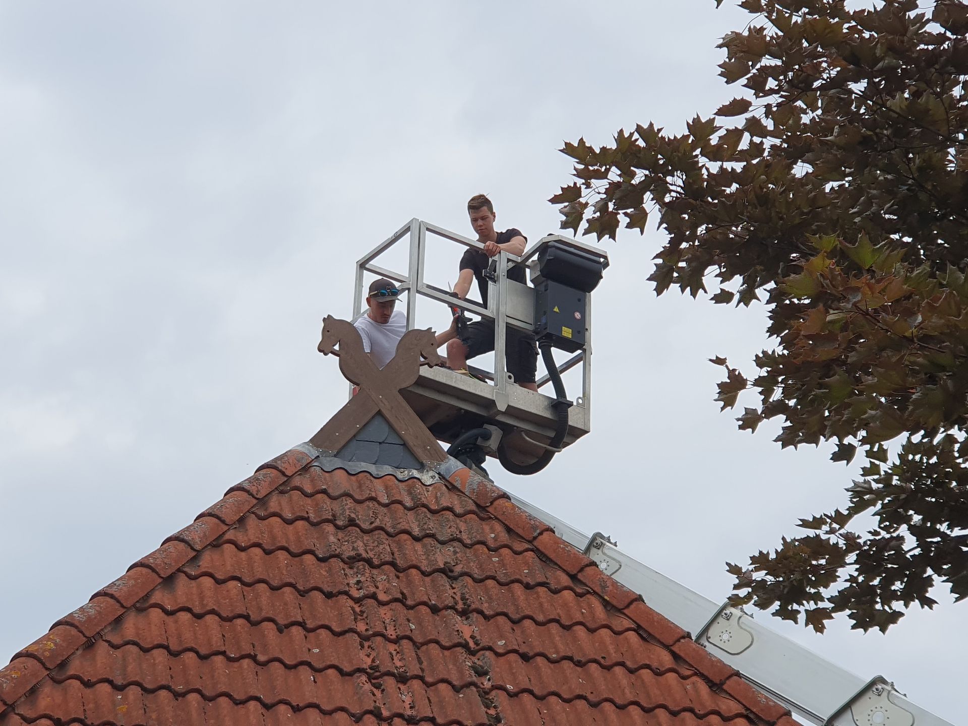 Zwei Arbeiter montieren auf einem Aufzug unter bewölktem Himmel eine Dachdekoration auf einem roten Ziegeldach.