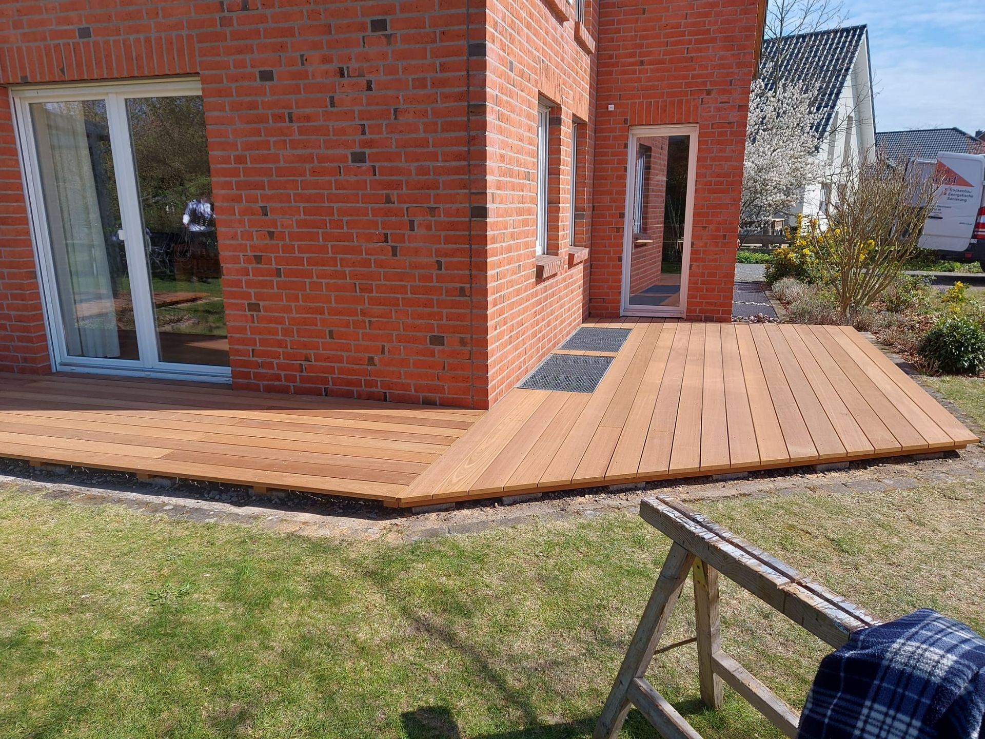 Holzterrasse vor einem roten Backsteinhaus, neben einem Rasenhof.