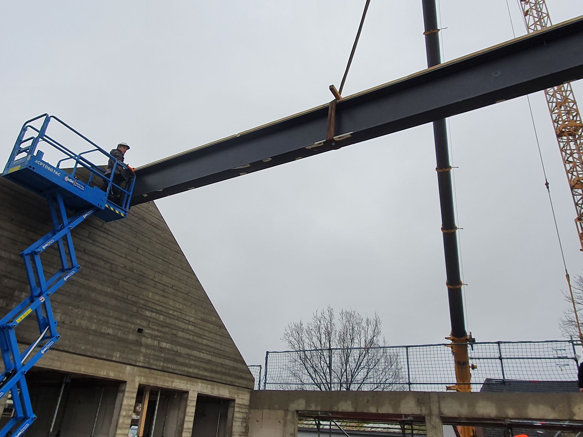 Bauarbeiter auf Hebebühne, die Stahlträger führt, der mit einem Kran abgesenkt wird.