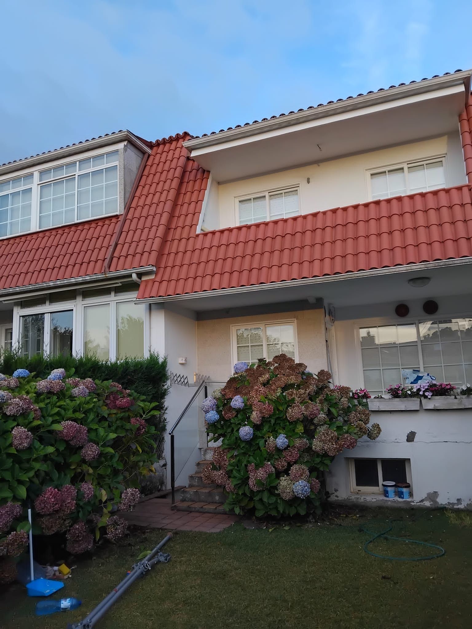 Casa de dos plantas con techo rojo, paredes blancas y arbustos en flor en el jardín delantero.