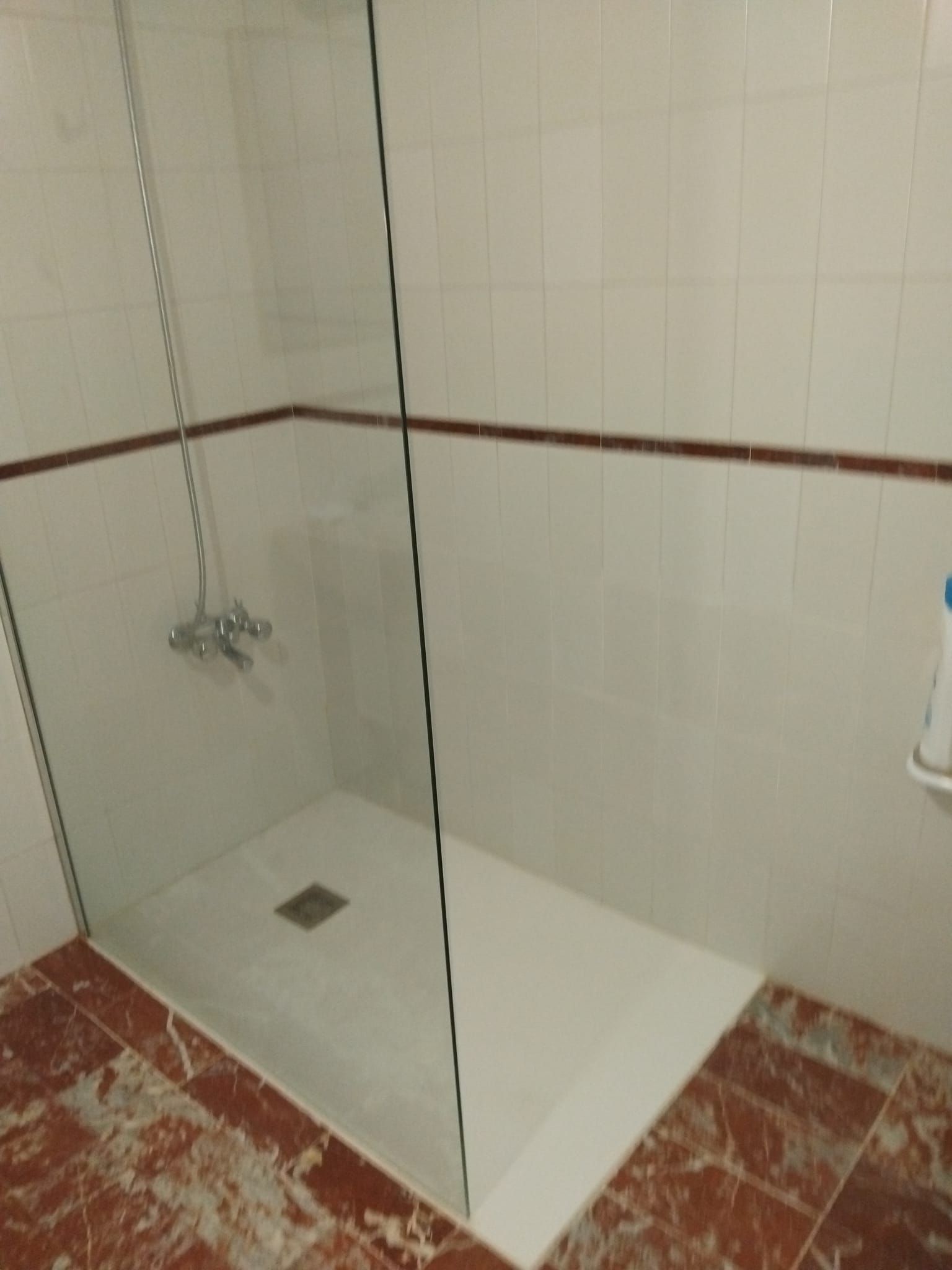 Cabina de ducha pequeña con azulejos, puerta de cristal, paredes blancas, suelo de baldosas rojas y desagüe en la esquina.