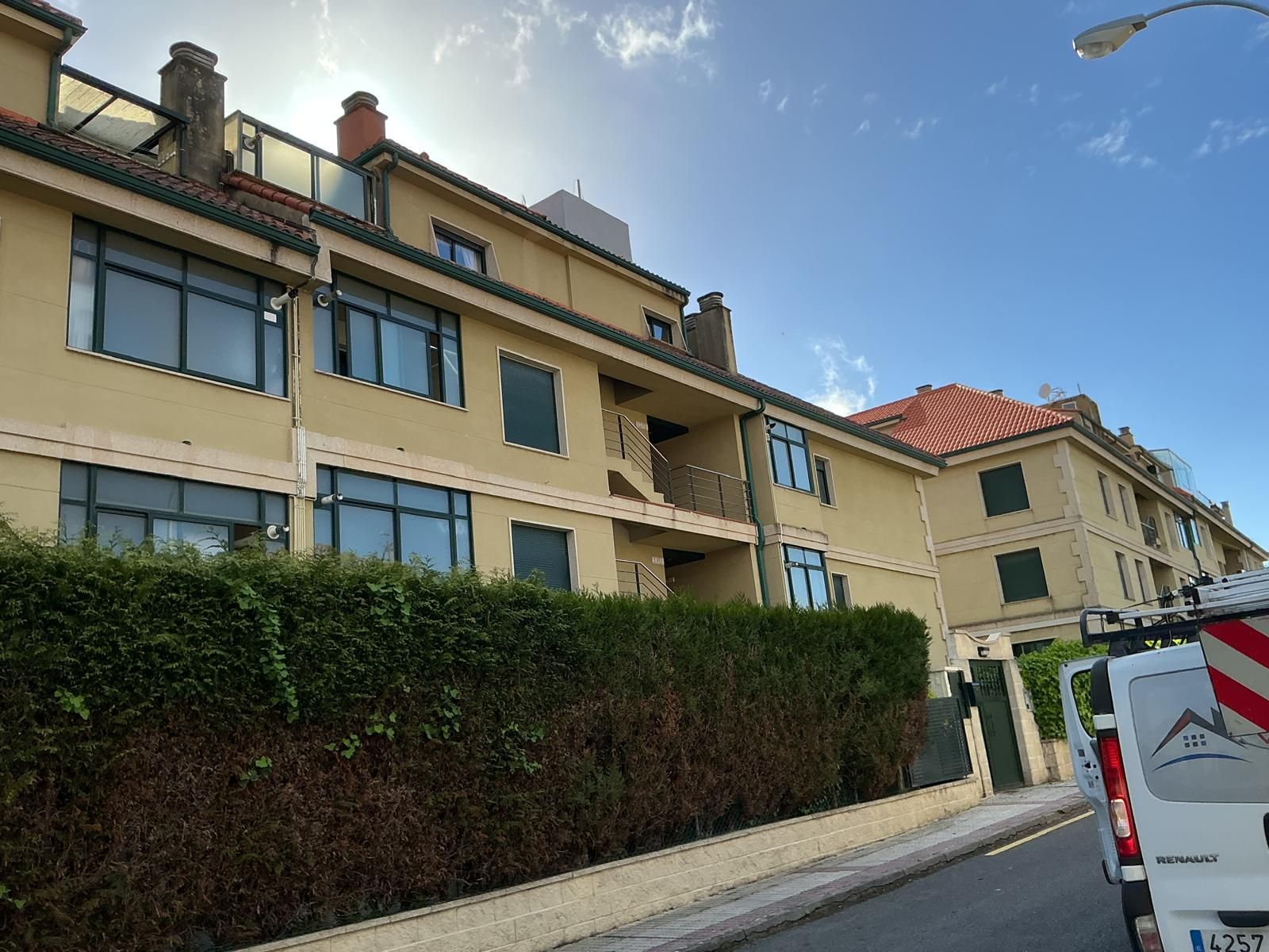 Calle residencial con edificios de apartamentos de color beige, tejados de tejas rojas y un seto bajo un cielo azul.