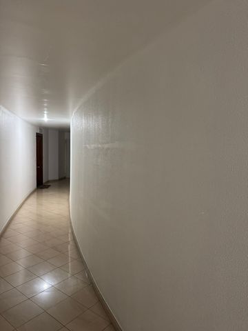Un estrecho pasillo blanco con una pared curva, suelo de baldosas beige y una puerta oscura al final.