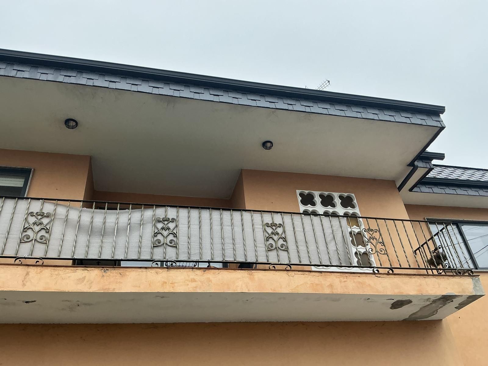 Vista desde un ángulo bajo del balcón de un apartamento beige con barandilla negra y techo blanco bajo un cielo gris.