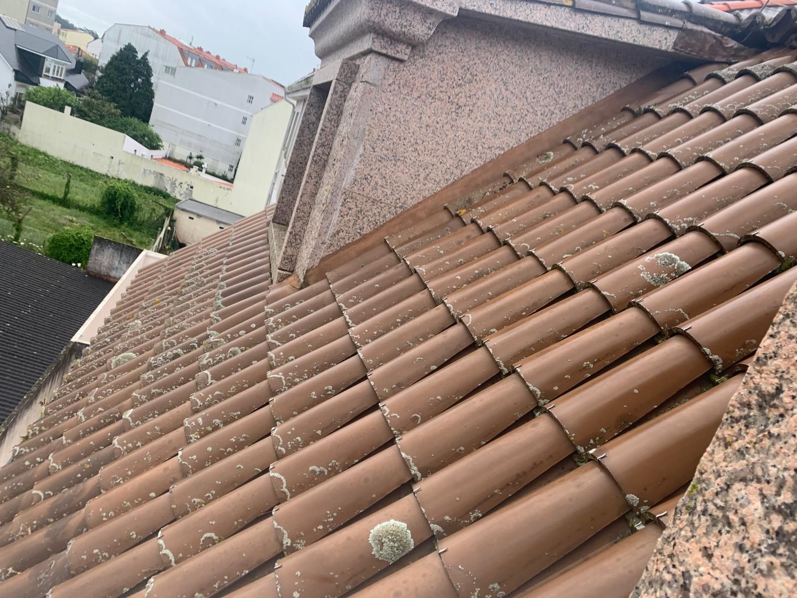 Tejado inclinado de tejas de terracota junto a una chimenea de estuco, con las casas vecinas visibles al fondo.