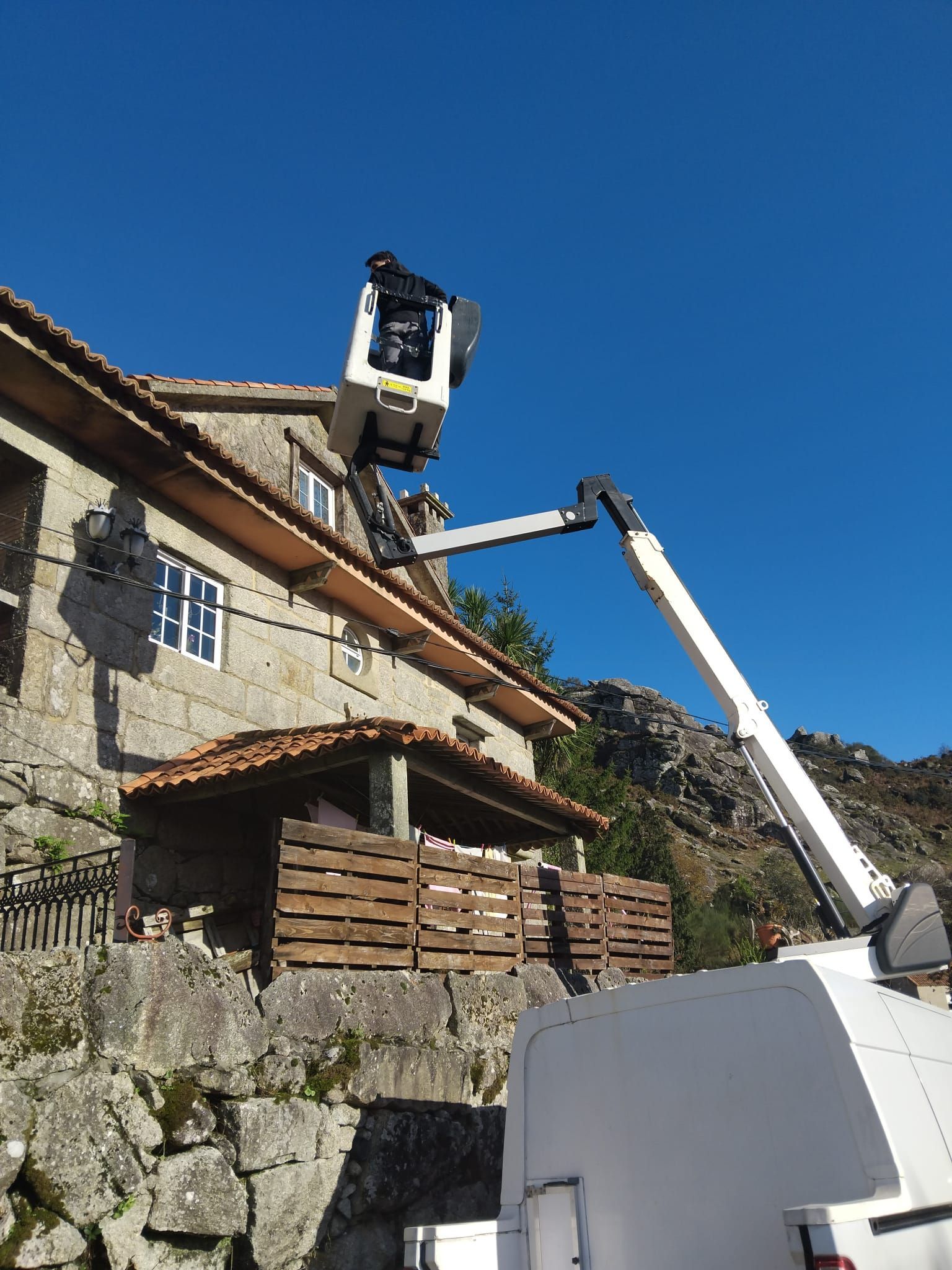 Un camión grúa eleva a un trabajador cerca de una casa de piedra en una ladera bajo un cielo azul despejado.