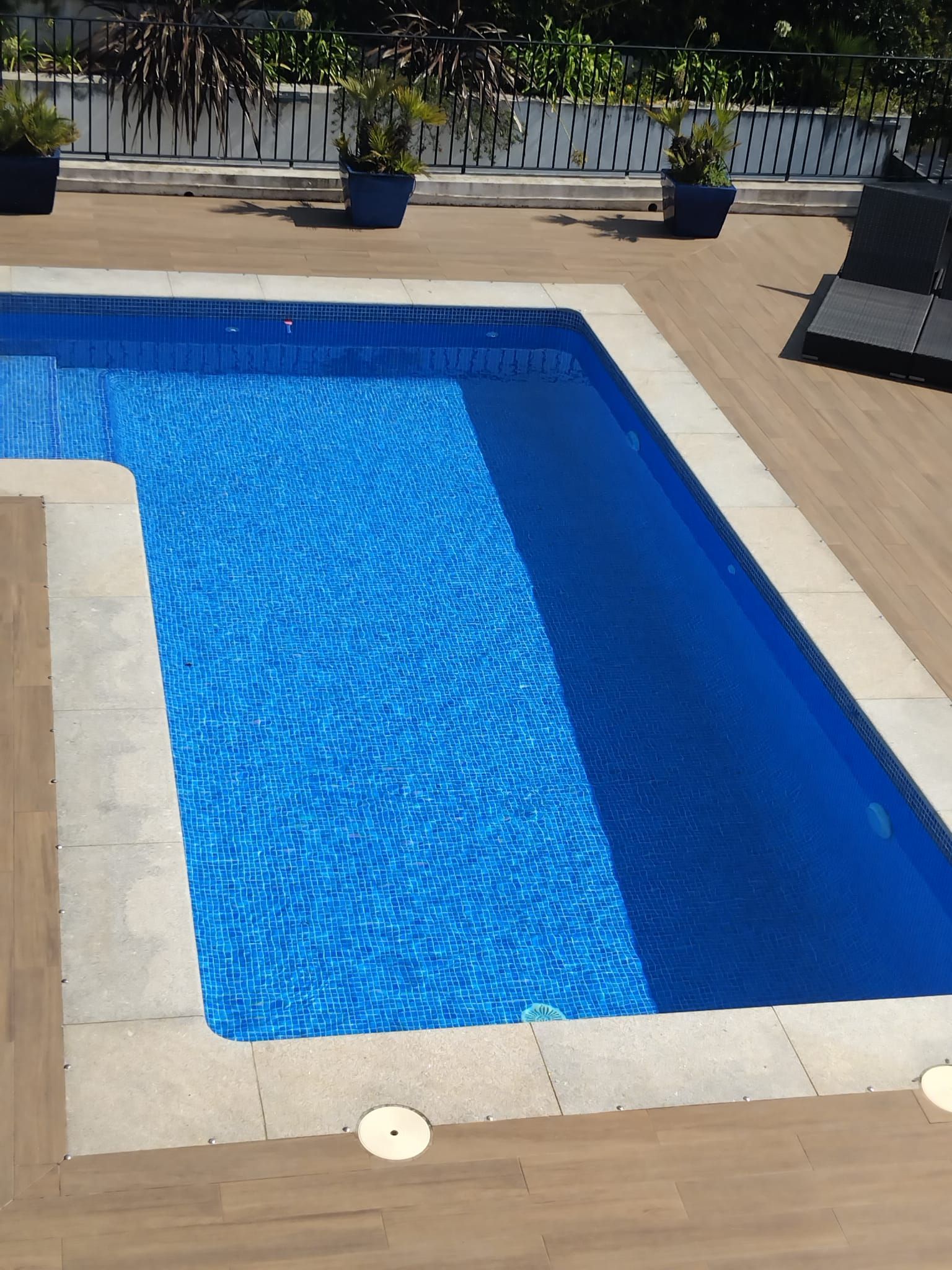 Piscina rectangular azul con terraza de piedra beige y plantas en macetas cerca.