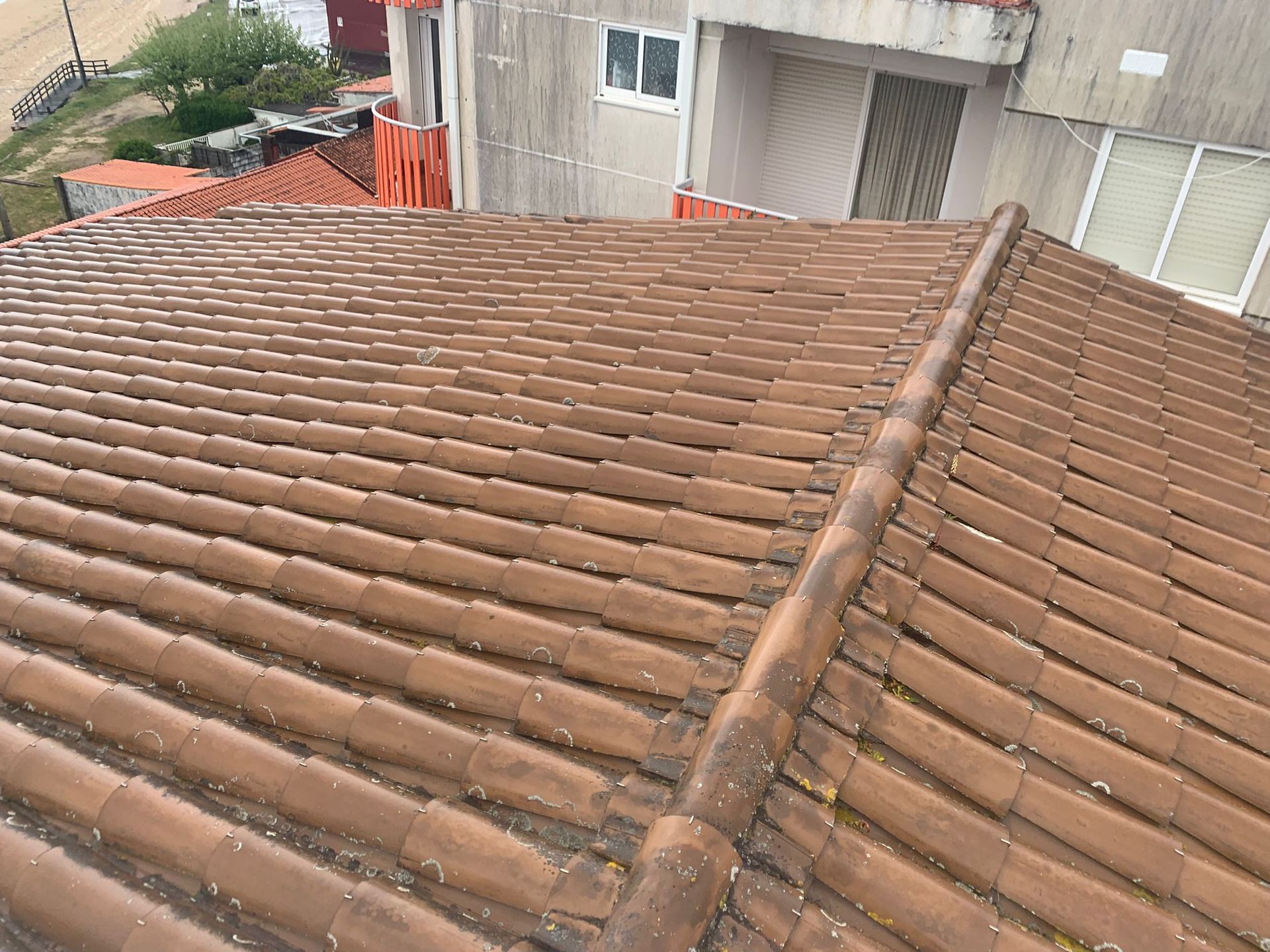 Tejado de tejas de terracota con cumbrera central, junto a un edificio de hormigón y una pequeña zona exterior.