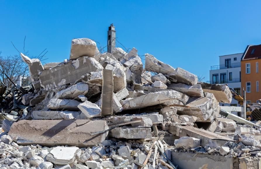 Montón de escombros de un edificio demolido bajo un cielo azul despejado, con una casa dañada al fondo.