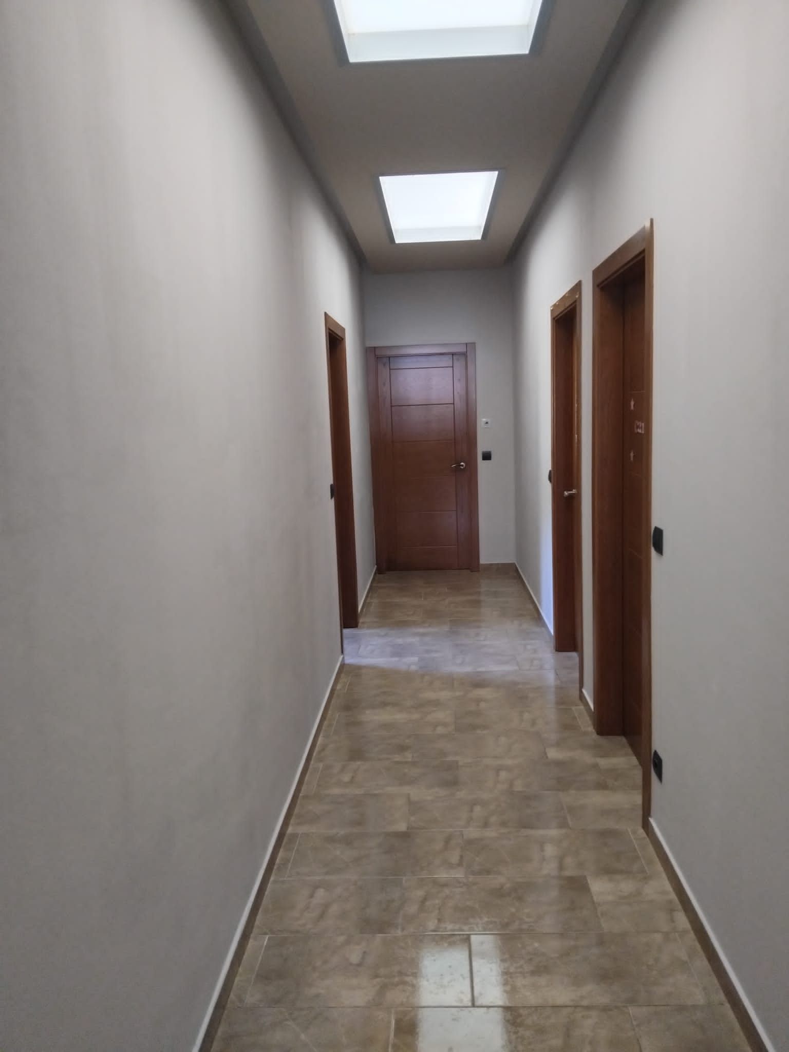 Pasillo estrecho con paredes beige, suelo de baldosas, puertas de madera y luces en el techo.