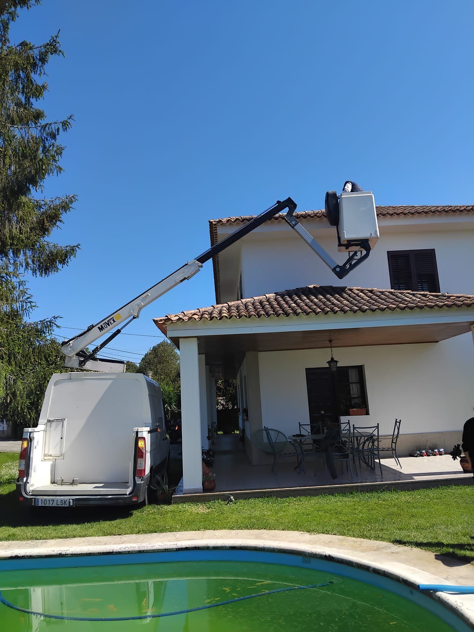 Camión utilitario blanco con plataforma elevadora que llega al tejado de una casa junto a una piscina en el patio