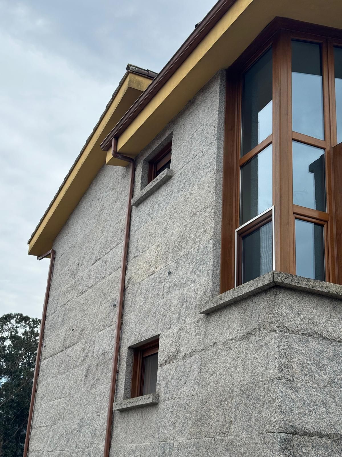 Esquina exterior de una casa de estuco con ventanas de marco de madera contra un cielo nublado.