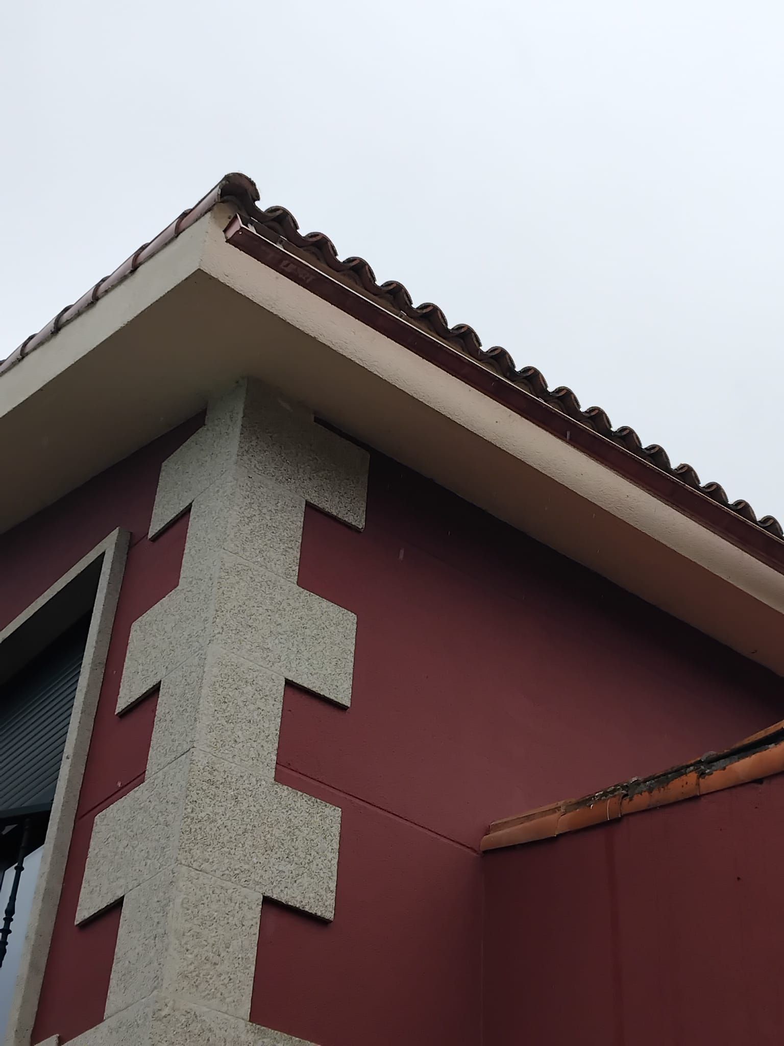 Esquina de una casa de estuco rojo con detalles de piedra y techo de tejas contra un cielo pálido.