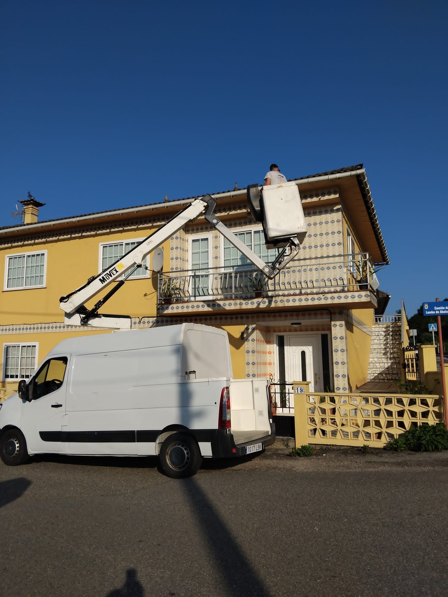 Un trabajador, subido a una plataforma elevadora, repara el tejado de una casa amarilla junto a una furgoneta blanca.