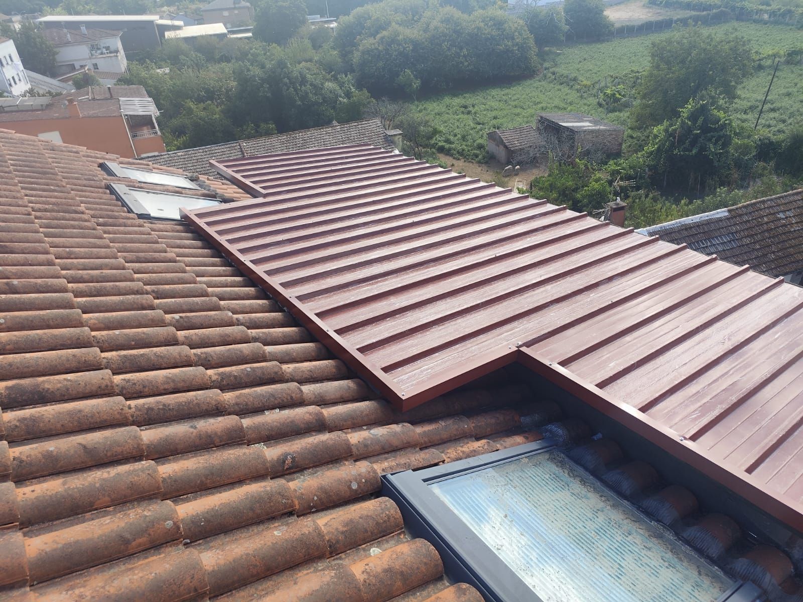 Azotea con tejas de terracota y un panel de techo de metal marrón, con vistas a campos verdes y árboles.