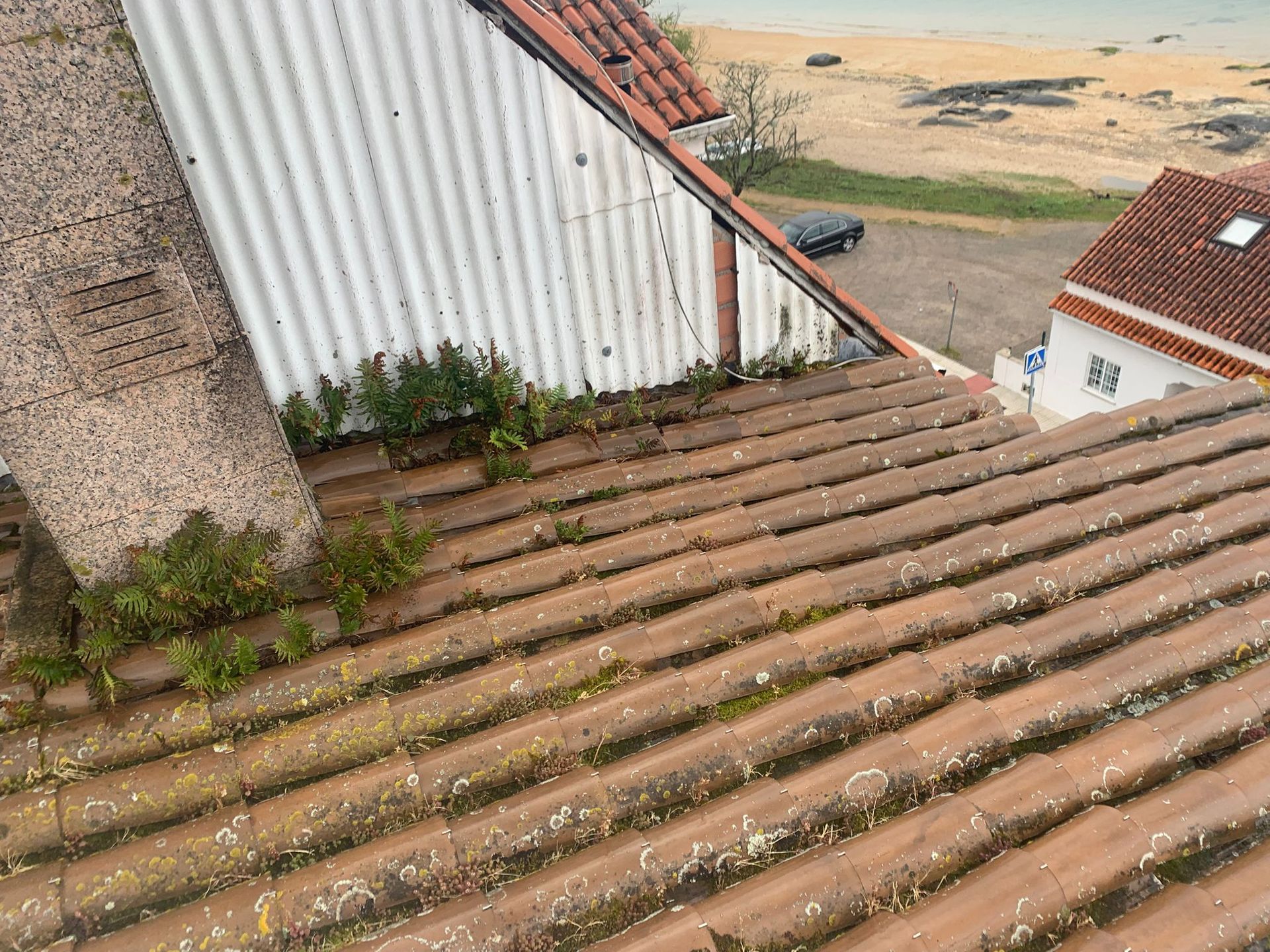 Tejado de tejas desgastadas junto a una pared blanca de chapa ondulada, con vistas a una playa de arena y a una carretera.