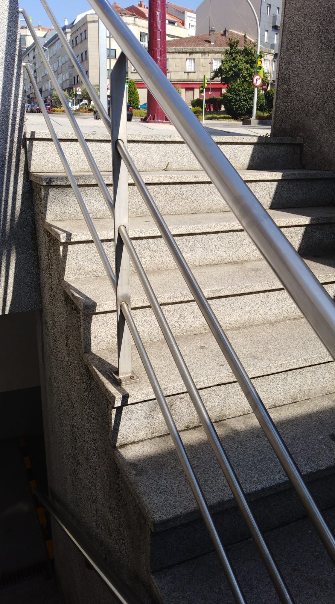 Barandilla metálica junto a unas escaleras exteriores de hormigón, bañadas por la luz del sol, con edificios al fondo.