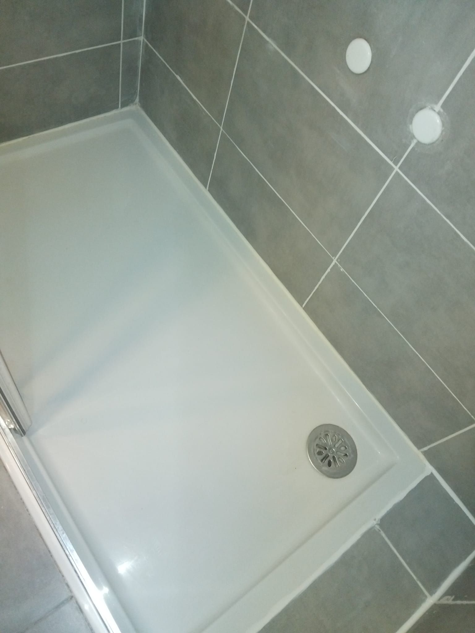 Plato de ducha blanco con desagüe en un rincón de baño con azulejos grises