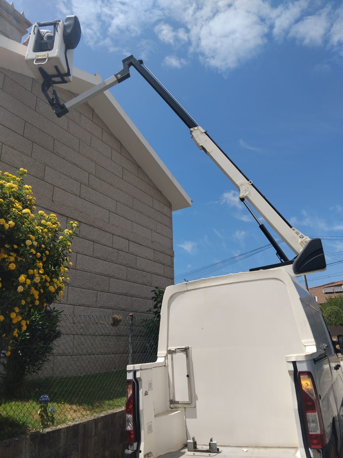 Camión grúa elevando a un trabajador en una plataforma elevadora junto al tejado de una casa bajo un cielo azul.