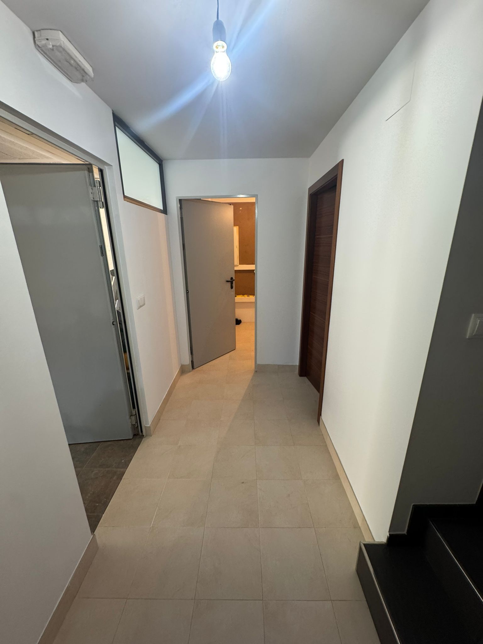 Pasillo estrecho con suelo de baldosas, paredes blancas, puertas abiertas y una sola bombilla colgante.
