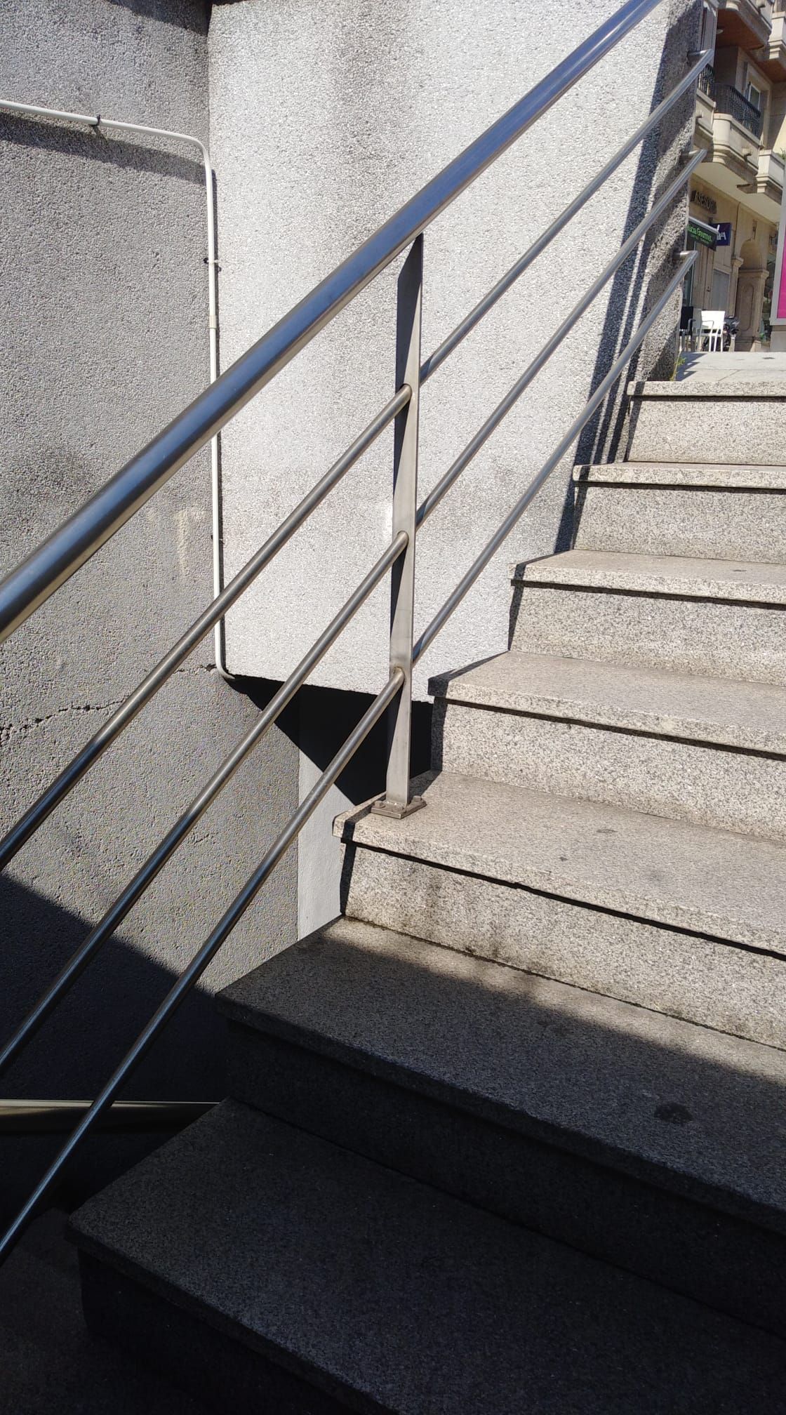 Escaleras de hormigón con barandilla de metal negro junto a una pared gris, iluminadas por la luz del sol y las sombras.