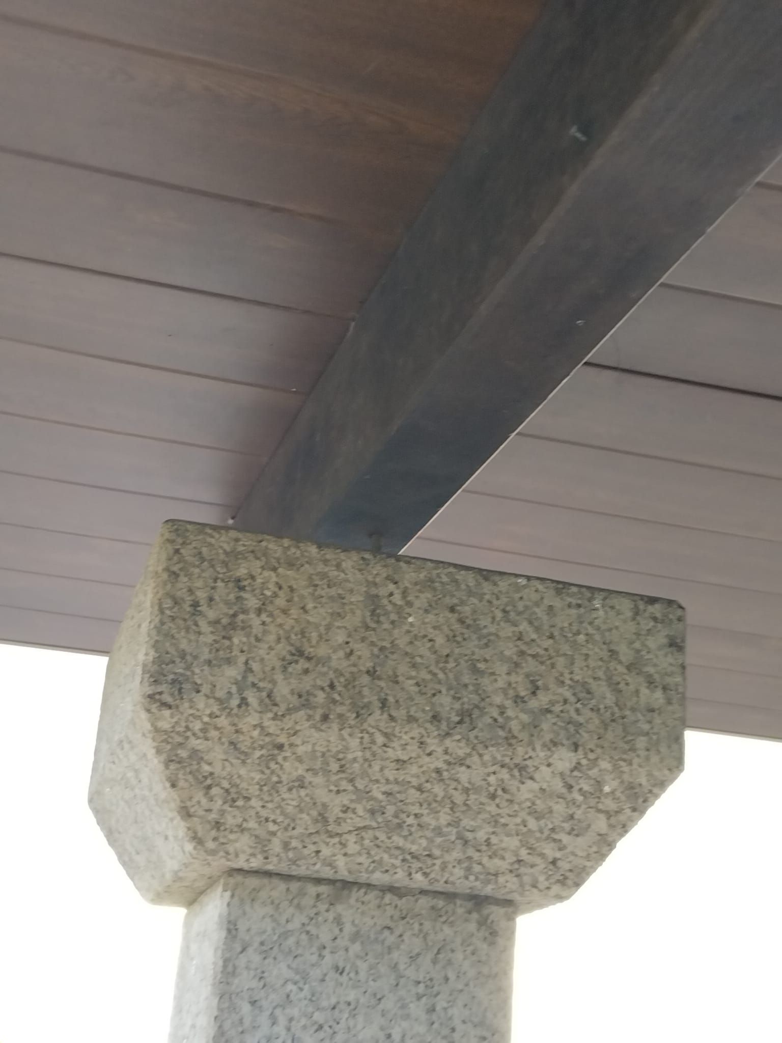 Pilar de hormigón que sostiene una viga de madera oscura bajo un techo cubierto.