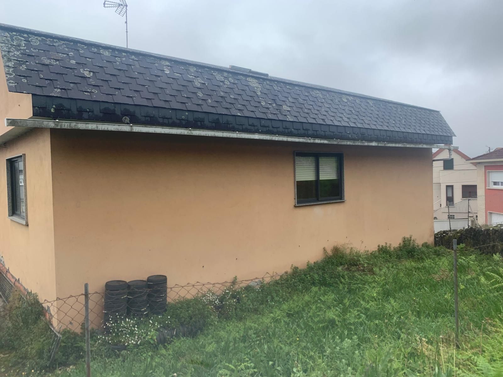 Pequeña casa beige con techo oscuro junto a la hierba crecida bajo un cielo nublado.