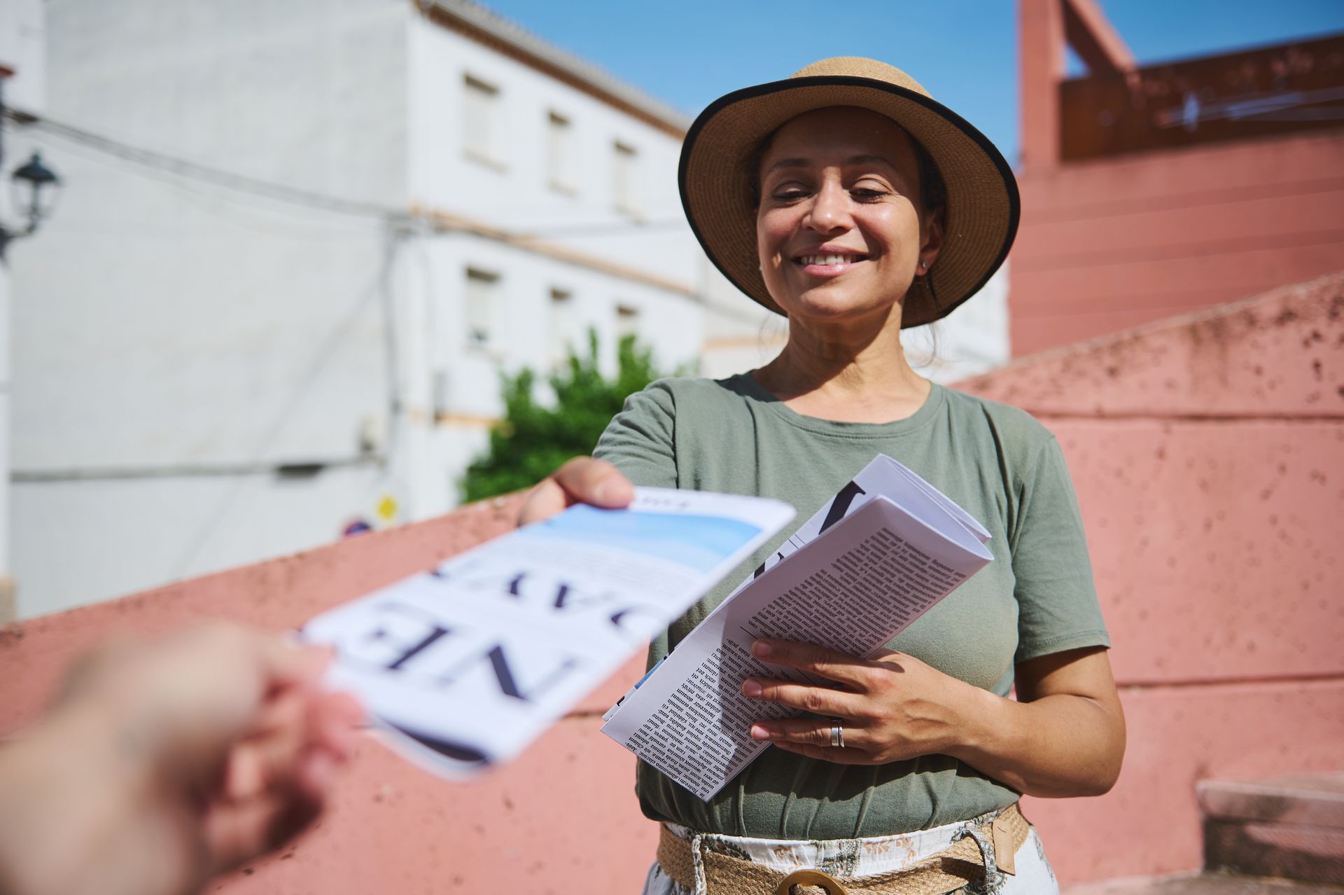 Une femme coiffée d'un chapeau distribue des brochures à une personne à l'extérieur, en souriant. Des bâtiments rose et blanc se dressent en arrière-plan.