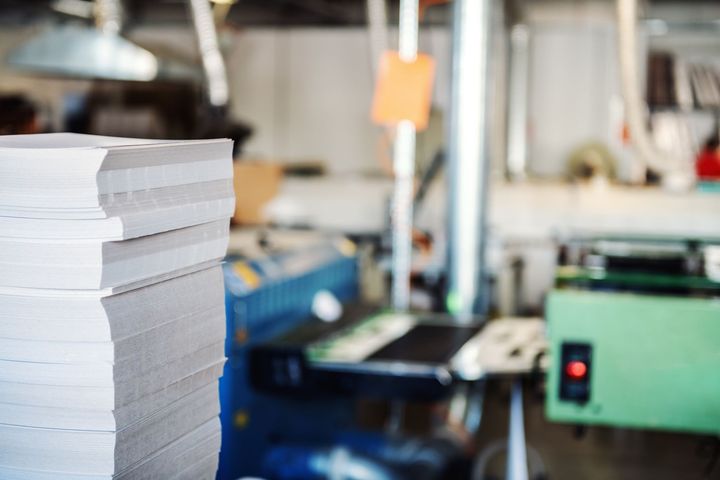 Pile de papiers blancs dans une imprimerie, machines floues en arrière-plan.