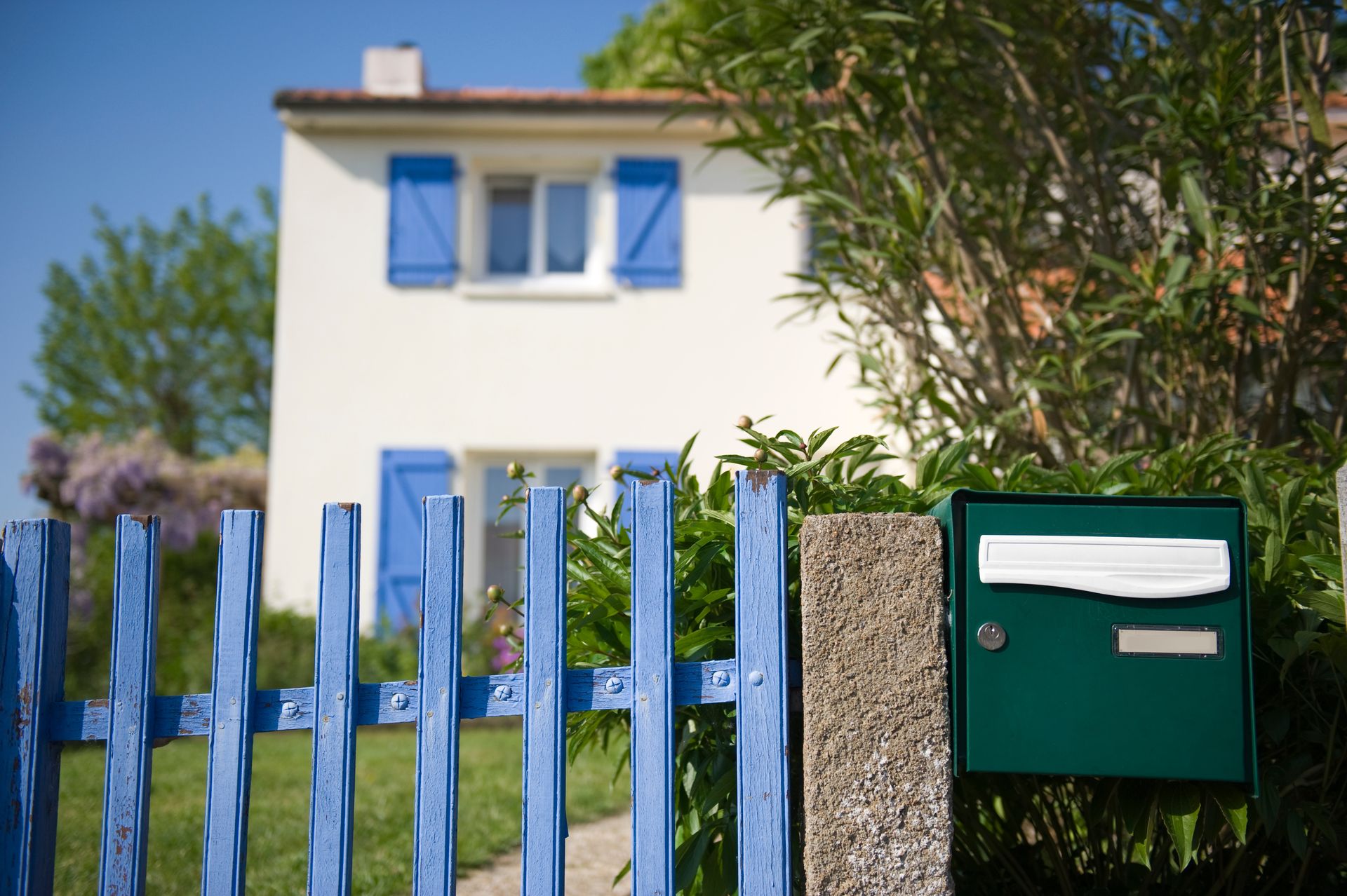 Clôture bleue, boîte aux lettres verte et maison blanche aux volets bleus par une journée ensoleillée.