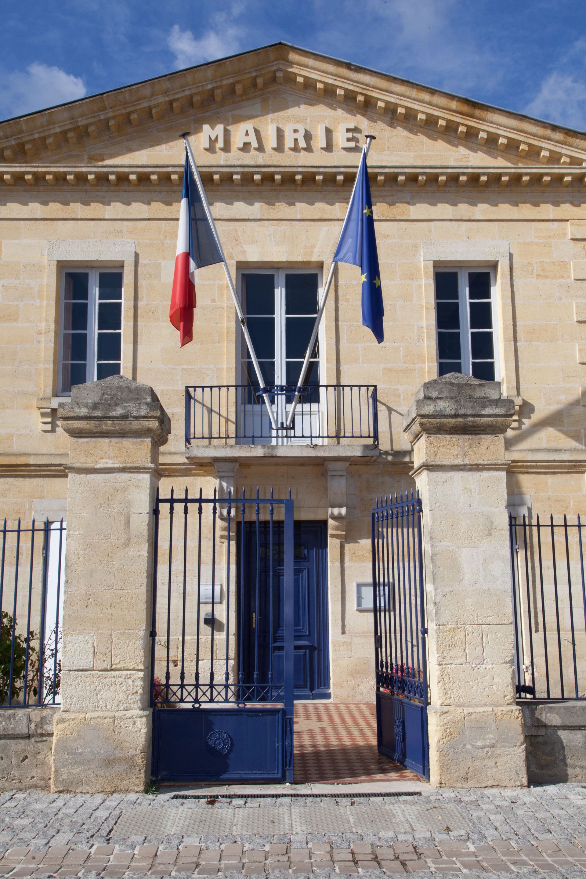 Hôtel de ville orné des drapeaux, français et européen. Portes bleues, fenêtres cintrées et façade en pierre claire.