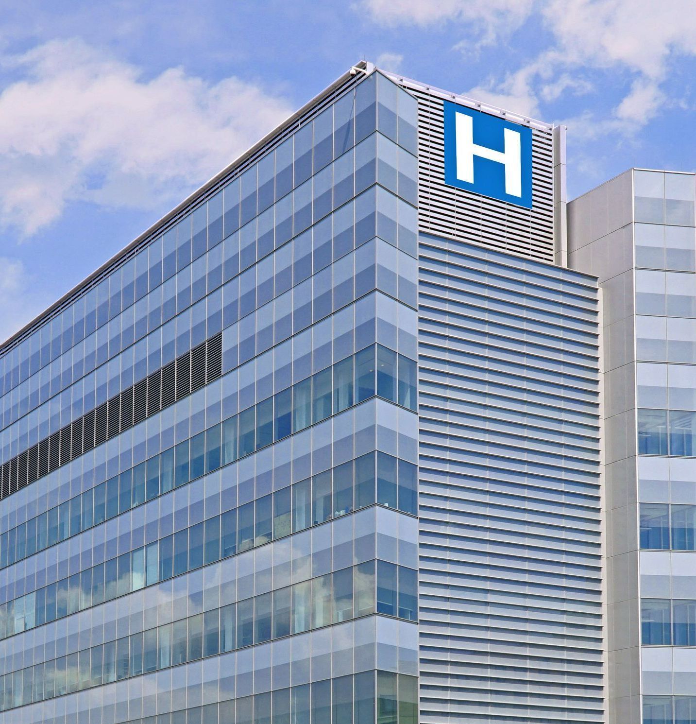 Bâtiment moderne en verre avec un panneau d'hôpital bleu indiquant la lettre H et ciel partiellement nuageux.