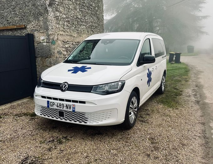 Ambulance blanche avec étoile de vie, stationnée sur un chemin en gravier, par temps couvert.