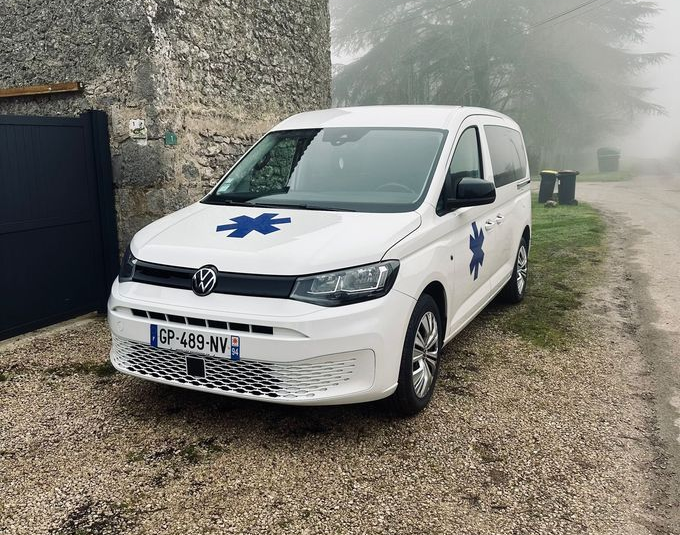 Ambulance blanche avec l'étoile bleue de vie ; garée à l'extérieur, logo VW visible.